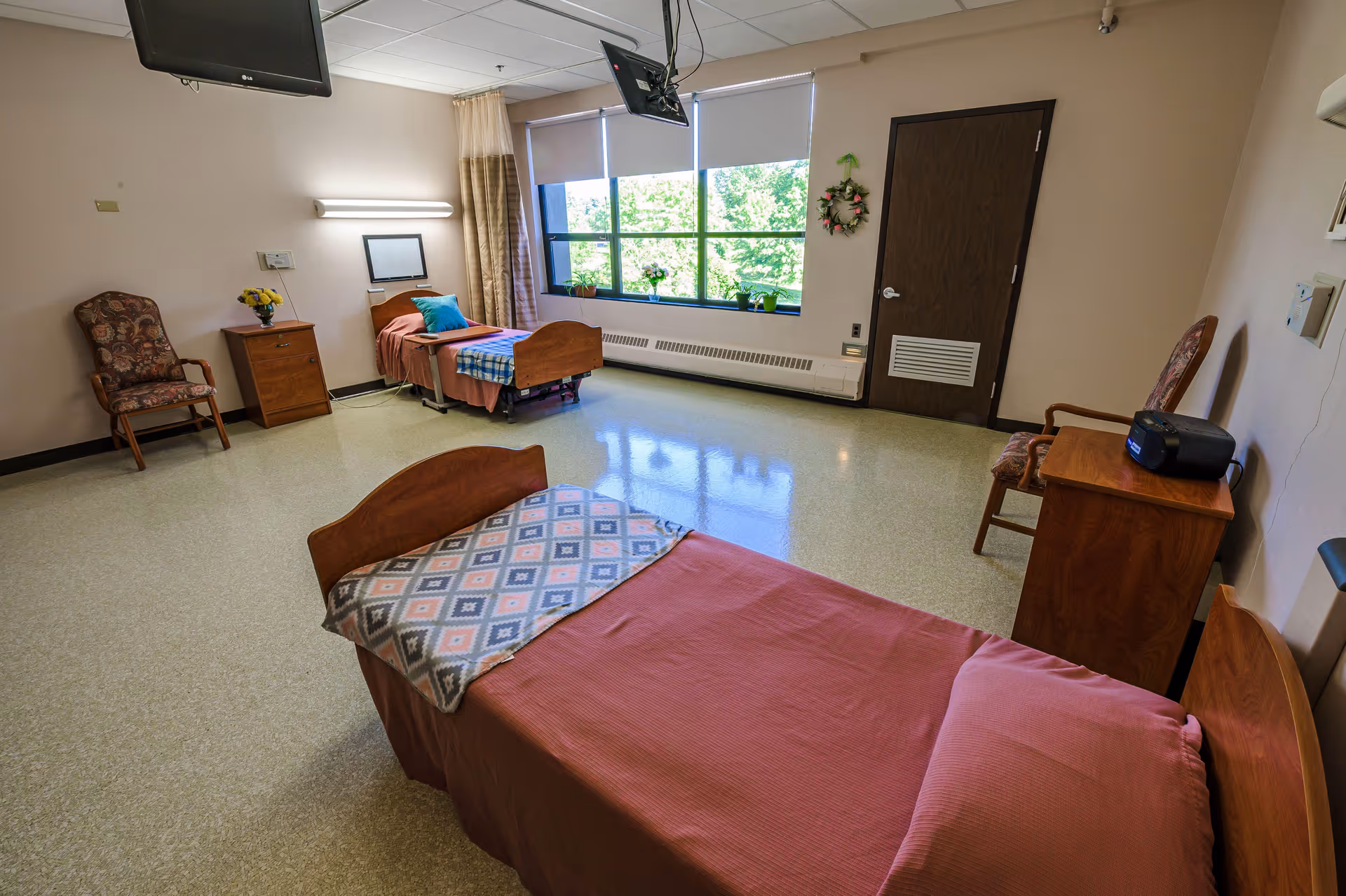 A spacious room in a senior living facility with two single beds, each with a pillow and blanket. The room has a large window with a view of green trees outside, two wooden chairs with patterned cushions, two wooden nightstands, a wall-mounted TV, and a small radio on one of the nightstands. The walls are light-colored, and the floor is covered with light-colored linoleum.