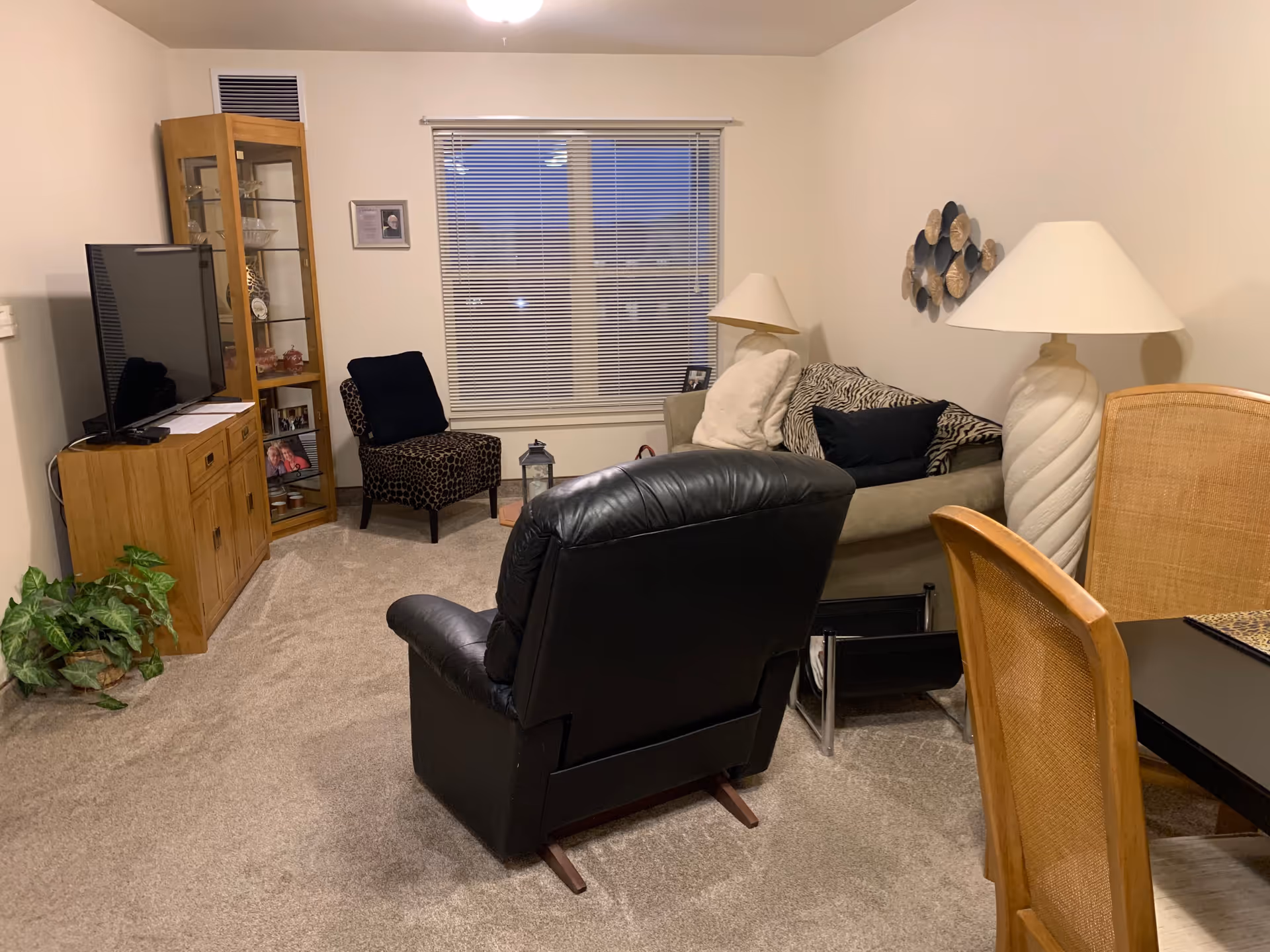 Living room with a black leather recliner facing a TV, a sofa with pillows, lamps, a glass display cabinet, dining chairs and a window with blinds.