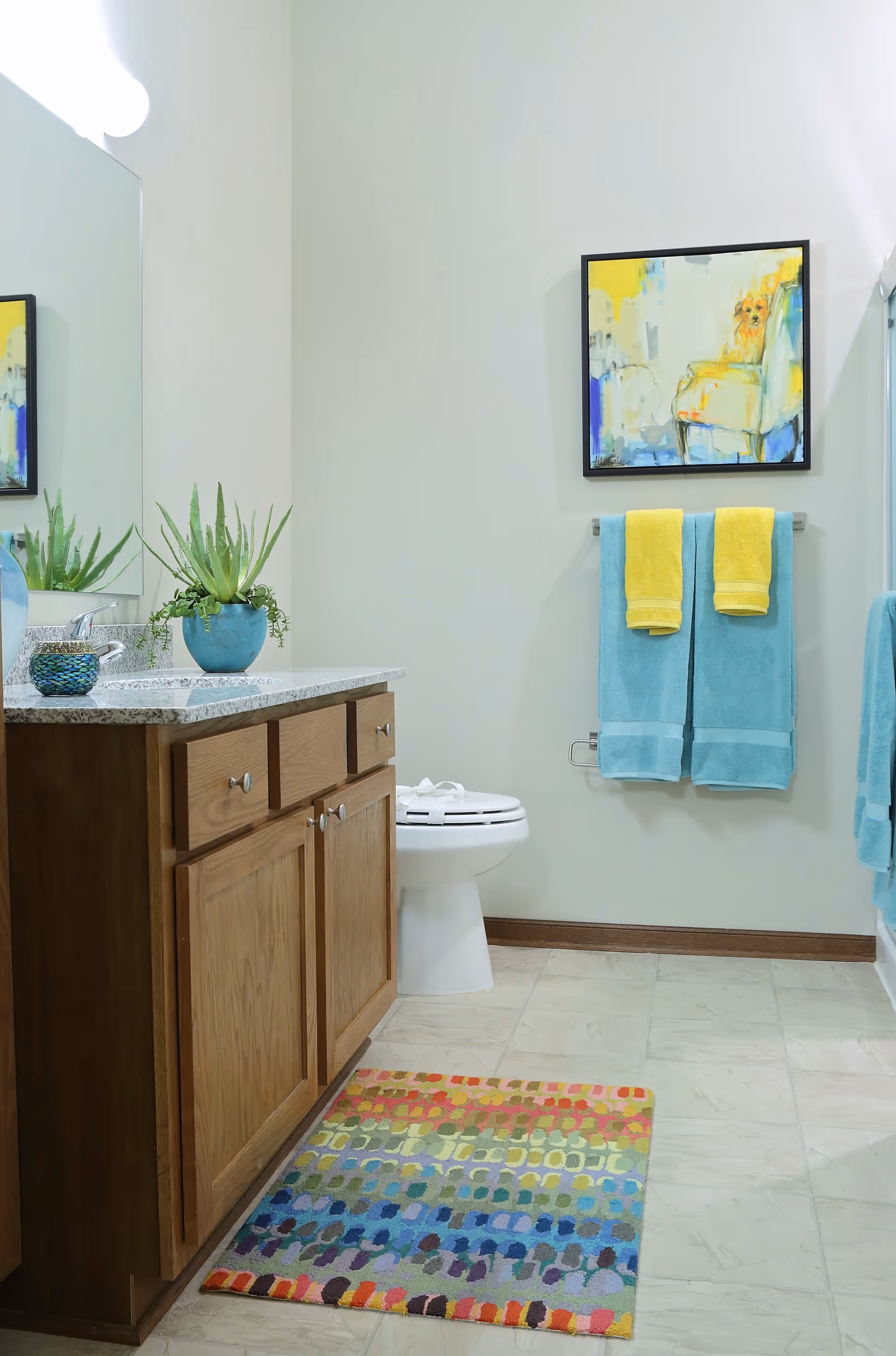 A clean and bright bathroom featuring a wooden vanity with a granite countertop, a potted green plant, a colorful patterned rug on the tiled floor, a white toilet, and two blue towels with yellow hand towels hanging on a wall-mounted towel rack. Above the towels is a framed abstract painting with yellow, blue, and white colors.