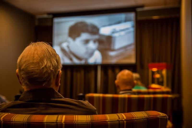 Several elderly individuals seated on striped couches in a dimly lit room watching a black and white movie projected on a screen.