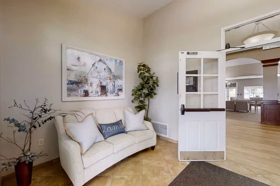 A cozy seating area with a cream-colored sofa adorned with three pillows, one of which has the word 'family' embroidered on it. A large framed painting of a barn hangs on the wall above the sofa. There are two potted plants, one on the left side and one behind the sofa. To the right, a white door with glass panels is open, leading into a spacious room with wooden flooring and additional seating visible in the background.