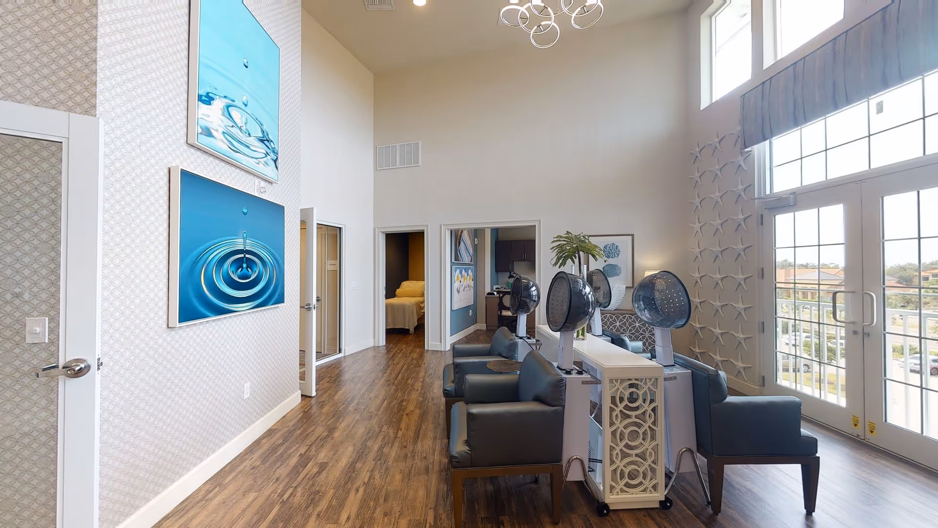 Interior view of a senior living facility room with high ceilings and large windows letting in natural light. The room features three black salon hair dryers with chairs, a white decorative console table, wooden flooring, and wall art depicting water droplets. There is a door leading to a bedroom with a bed visible and another door leading to a kitchenette area. The wall near the windows is decorated with starfish ornaments.