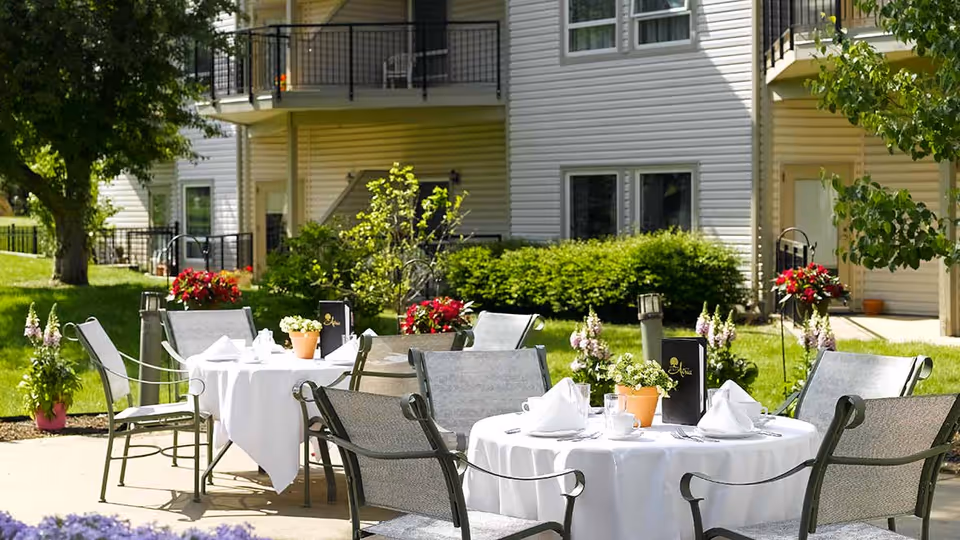 Outdoor patio area at Celebration Villa of Hearthstone with round tables covered in white tablecloths, set with napkins, cups, and small potted plants. Metal chairs surround the tables, and there are flowers and greenery in the background near a multi-story building with balconies.