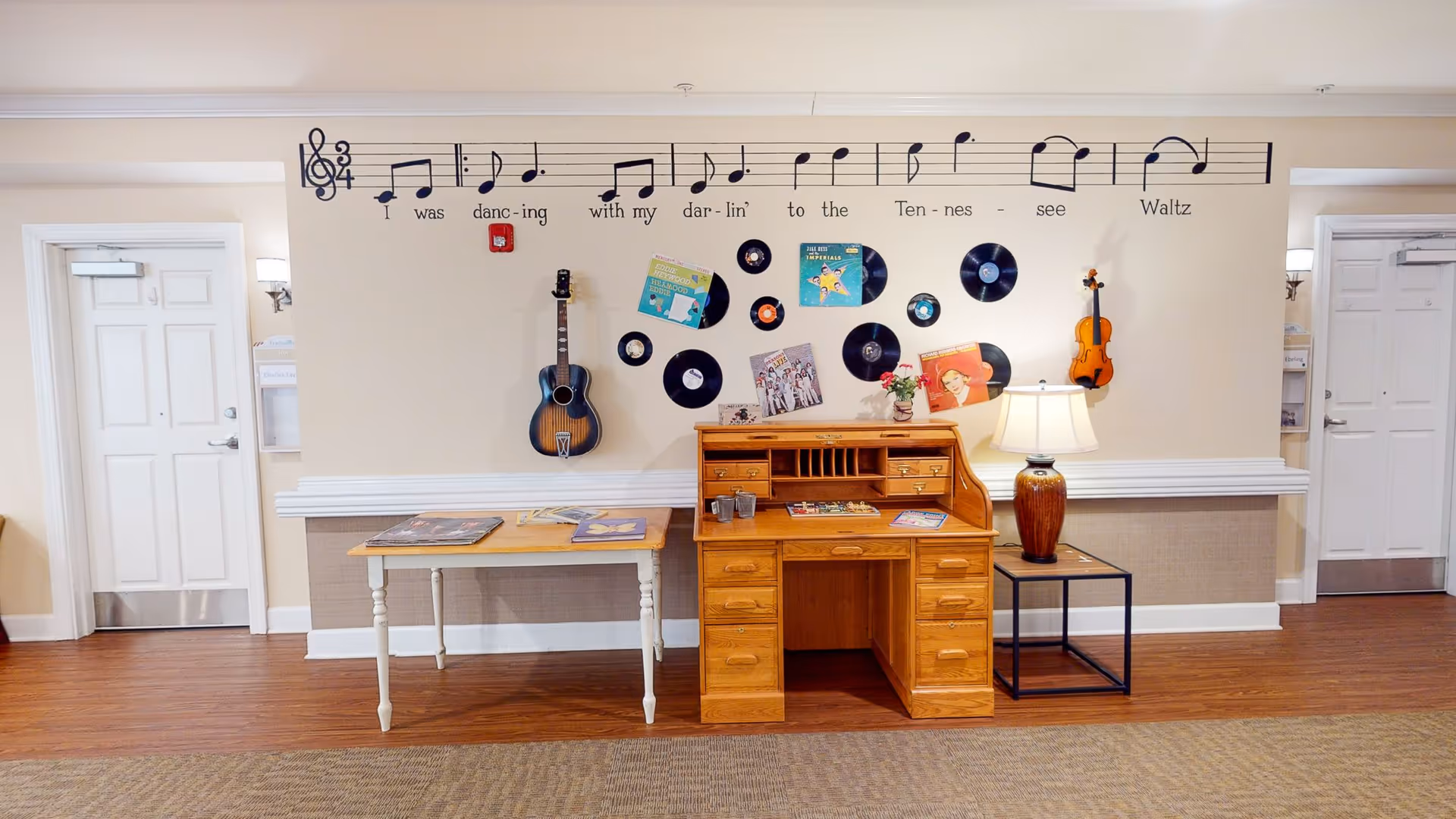 A hallway area in a senior living facility with musical-themed wall decorations including a large musical staff with notes and lyrics, vinyl records, and a guitar and violin mounted on the wall. Below the decorations is a wooden roll-top desk with a table lamp on a small side table next to it, and a small table with books or magazines. Two white doors are visible on either side of the wall.