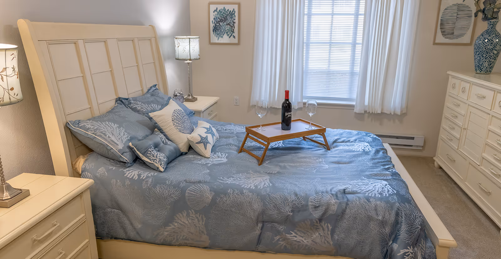 A cozy bedroom with a white wooden bed frame and blue bedding featuring coral and seashell patterns. There are several decorative pillows on the bed, and a wooden tray with a bottle of red wine and two wine glasses. The room has two nightstands with lamps, a white dresser with a blue vase, and two framed artworks on the walls. A window with white curtains allows natural light into the room.