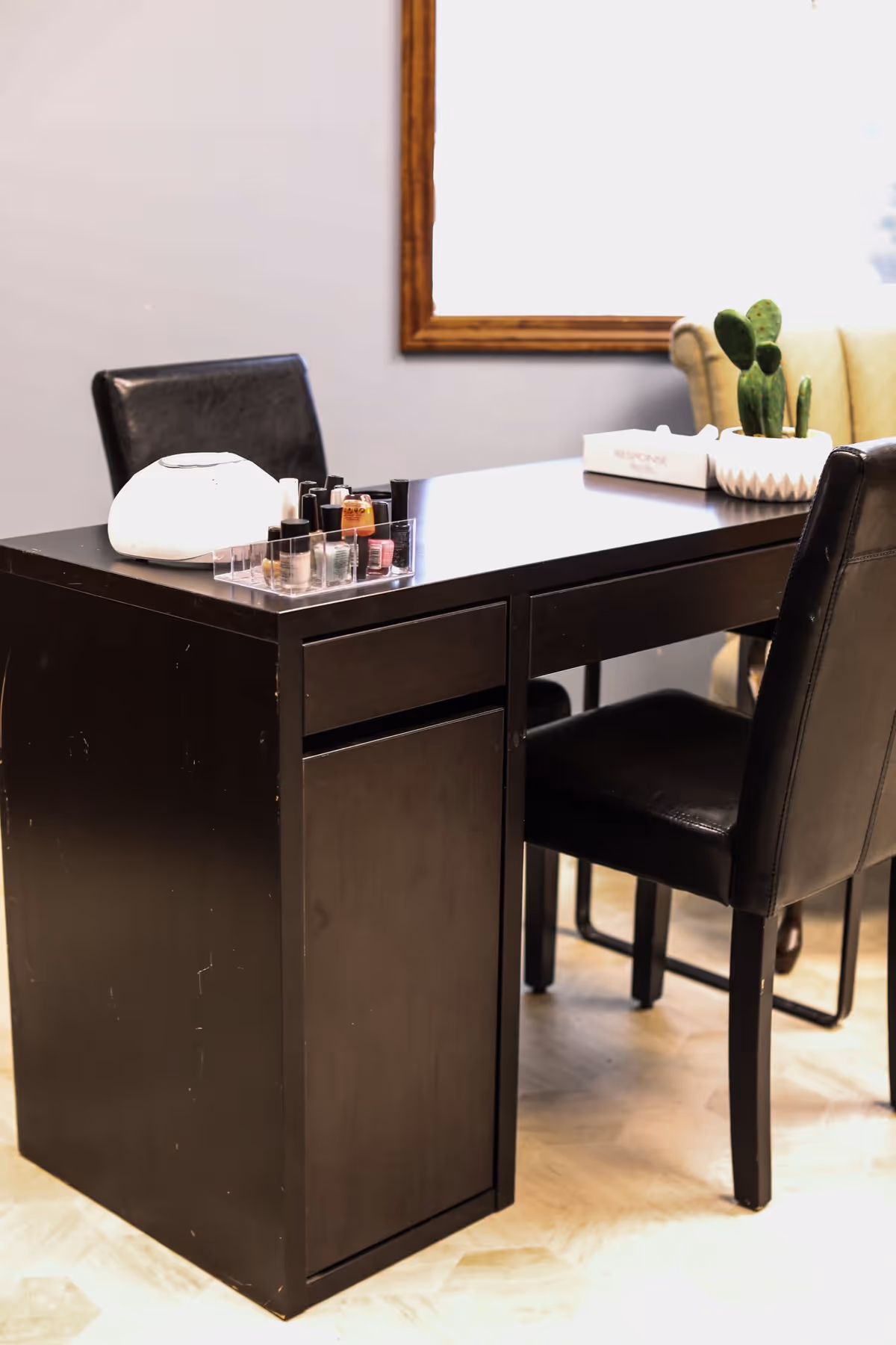 A dark wooden table with two black leather chairs. On the table, there is a white round object, a clear organizer holding various nail polish bottles, and a white planter with a small cactus. A beige upholstered chair and a window with a wooden frame are visible in the background.