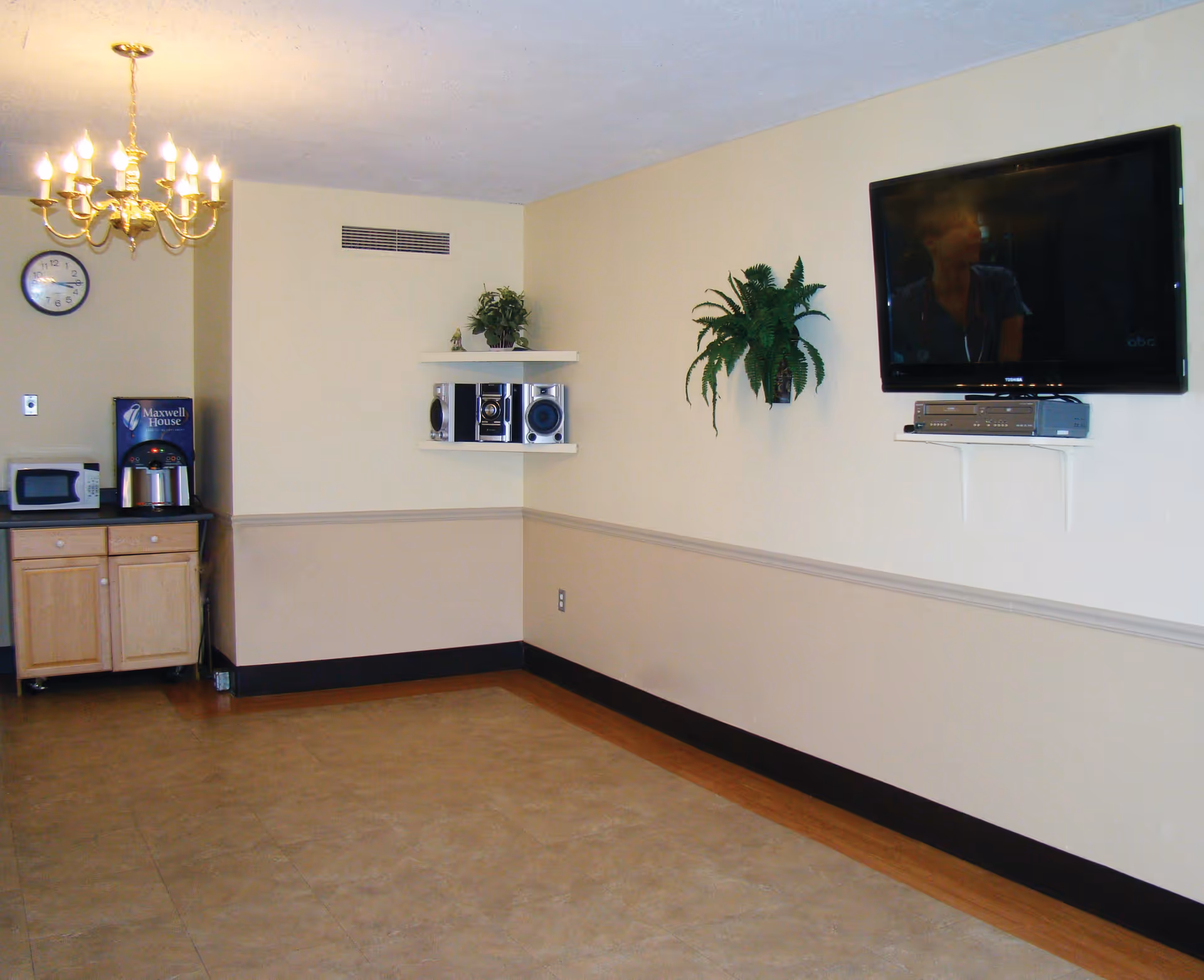 A small common area with a mounted flat-screen TV, a shelf with a stereo system, a hanging plant, and a corner kitchenette featuring a microwave, coffee maker, and a clock on the wall under a chandelier.