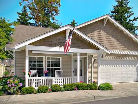 A beige cottage with white trim, front porch seating, and an American flag.