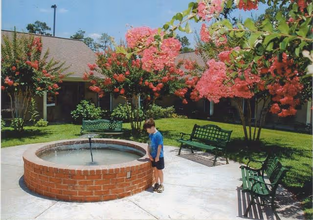 A young boy stands near a circular brick water fountain in a courtyard with green benches and blooming pink flowering trees. The courtyard is surrounded by a building with a sloped roof and windows.