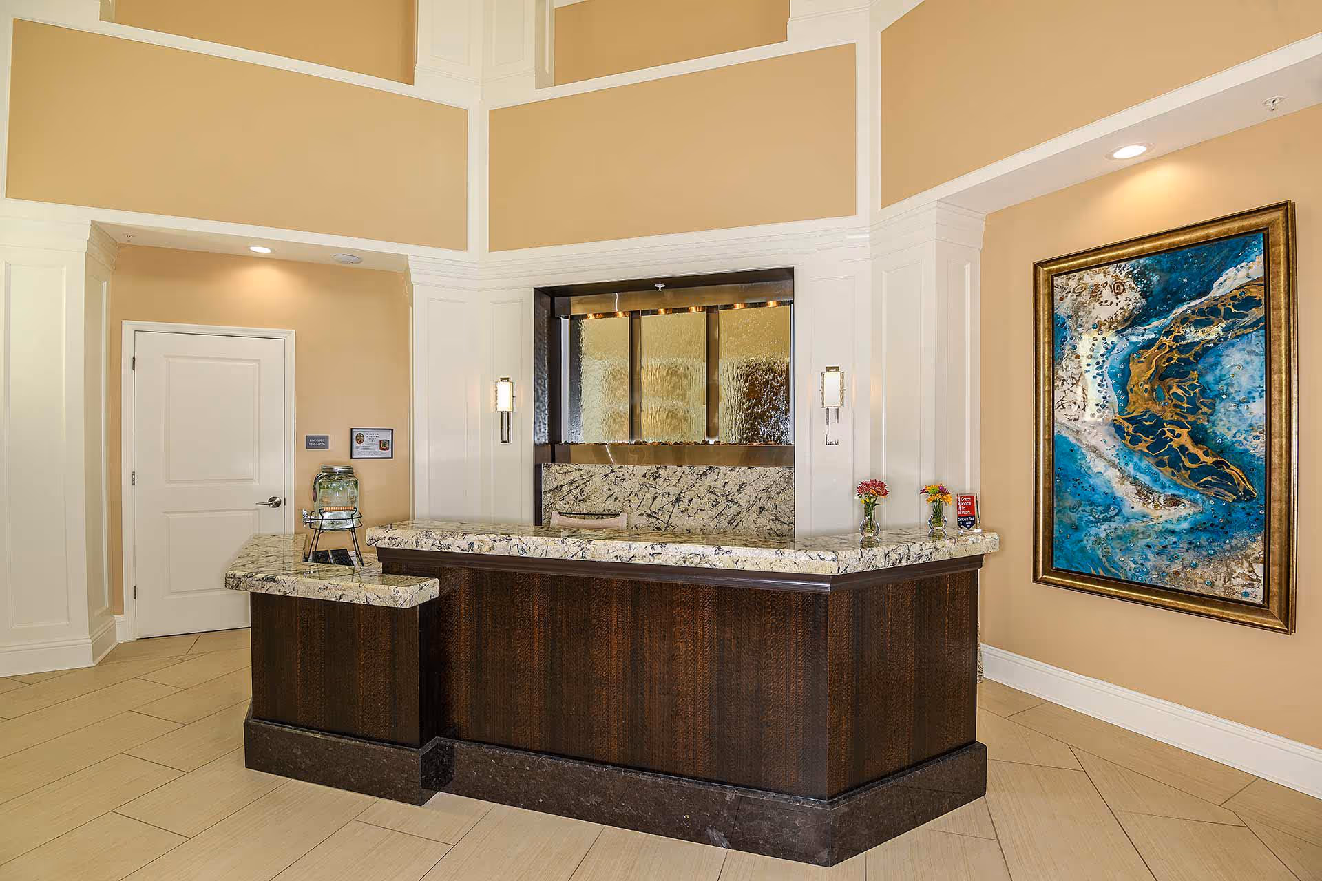 Reception area with a dark wood and marble counter, beige walls, a door on the left, a large abstract blue and gold painting on the right wall, and two small flower vases on the counter.