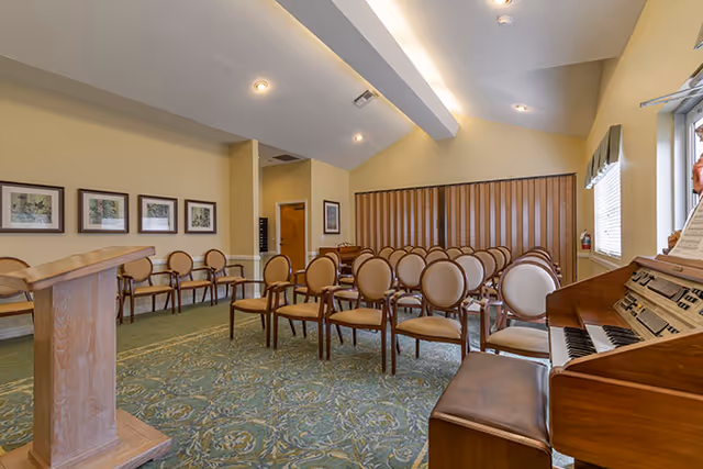 Interior view of a meeting or chapel room with rows of beige cushioned chairs arranged facing a wooden podium. There is a wooden organ with sheet music on the right side near a window with blinds. The room has framed pictures on the left wall and a patterned carpet on the floor.
