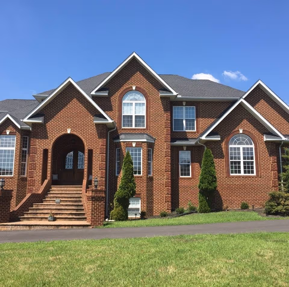 Front exterior view of a large brick building with multiple gables, arched windows, and a central entrance with steps leading up to double wooden doors. The building is surrounded by a well-maintained lawn and small shrubs under a clear blue sky.