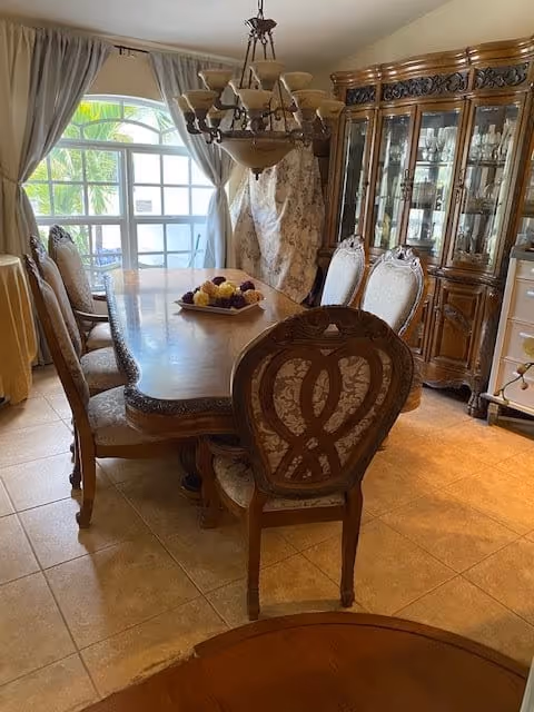 A dining room with a wooden dining table surrounded by eight ornate chairs with patterned upholstery. A chandelier hangs above the table. There is a large wooden china cabinet with glass doors displaying glassware on the right side. A window with curtains allows natural light into the room.