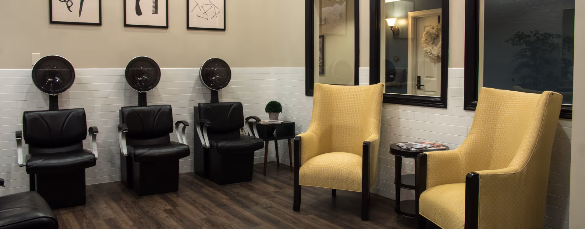 Interior of a salon area with three black hair dryer chairs lined up against a wall with white subway tiles and three framed abstract art pieces above. To the right, there are two yellow upholstered armchairs separated by a small round table with magazines on it. The floor is dark wood, and there are large windows with black frames on the right wall.