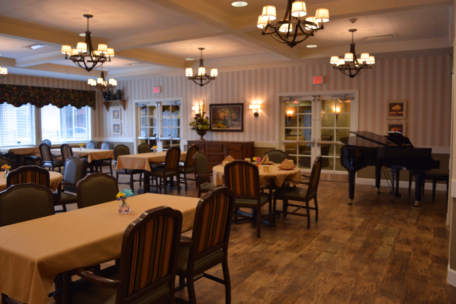 A spacious dining room with multiple tables covered in beige tablecloths, each surrounded by chairs with striped upholstery. The room features wooden flooring, several chandeliers with warm lighting, wall sconces, framed artwork, and a black grand piano in the corner. Large windows with blinds and floral valances allow natural light to enter the room.