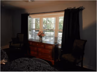 Bedroom with a wooden chest of drawers topped with a vase of flowers, flanked by two chairs beneath a bay window with dark curtains.