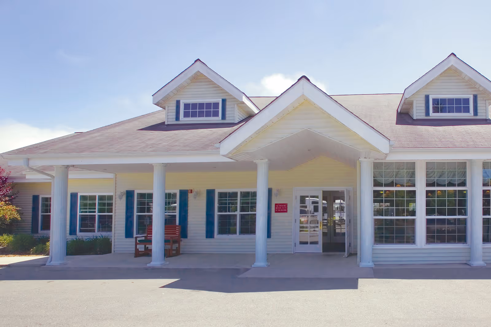 Front exterior view of a single-story building with white siding, blue window shutters, and a covered entrance supported by four white columns. There are two dormer windows on the roof and a bench near the entrance.