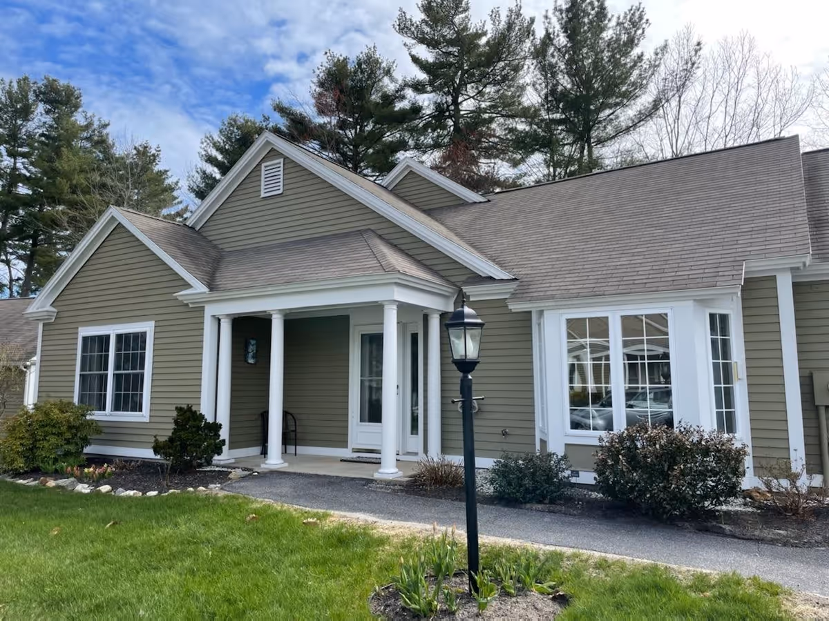 Exterior view of a single-story residential building with green siding, white trim, and a covered porch supported by white columns. There is a black lamp post in front of the building, surrounded by green grass, shrubs, and trees in the background under a partly cloudy sky.