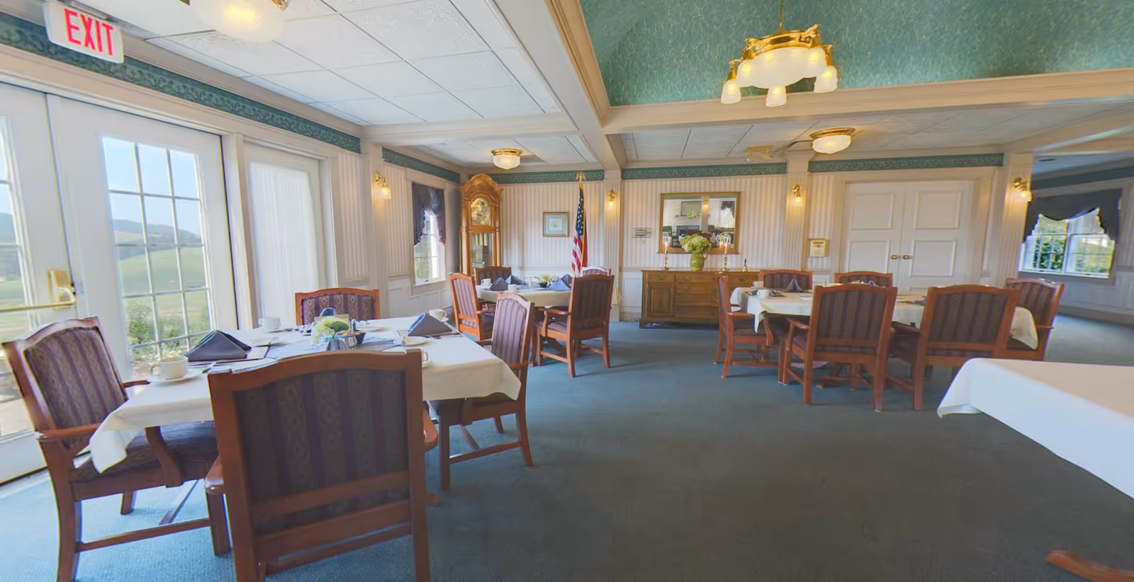 A dining room with several tables covered with white tablecloths and set with cups, napkins, and small flower arrangements. The room has wooden chairs with patterned upholstery, a grandfather clock, an American flag, and a sideboard with a mirror and decorative items. Large windows and glass doors allow natural light to enter, and the ceiling has decorative beams and light fixtures.