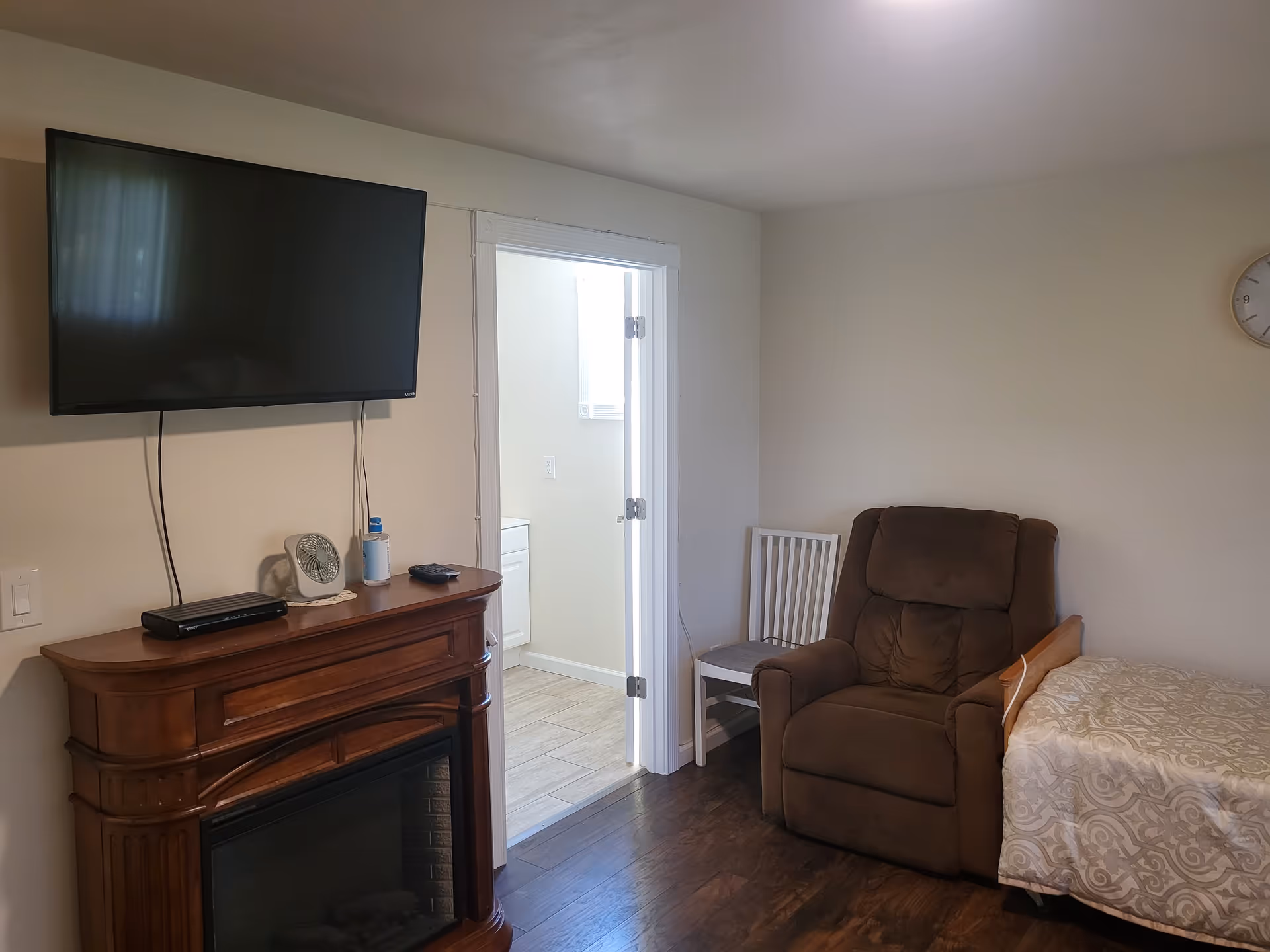 A cozy room with a brown recliner chair, a white wooden chair, and a single bed with a patterned beige and white bedspread. A wooden electric fireplace with a flat-screen TV mounted above it is on the left wall. There is an open door leading to a bright bathroom with tiled flooring.