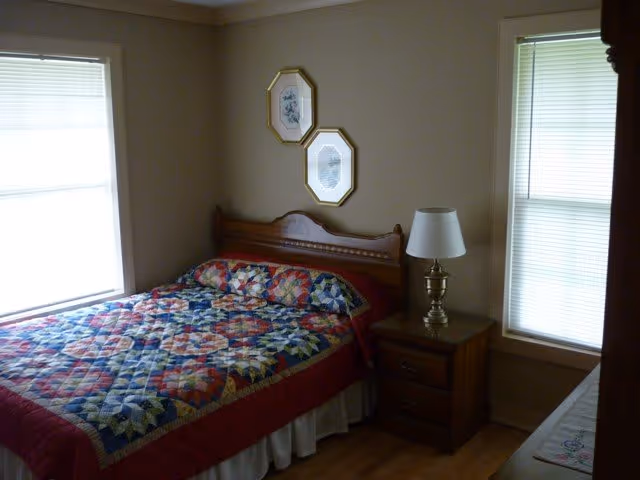 A bedroom with a wooden bed frame and a colorful patchwork quilt on the bed. There are two framed pictures hanging on the wall above the headboard. A wooden nightstand with a brass lamp is positioned next to the bed. The room has two windows with closed blinds, and wooden flooring.