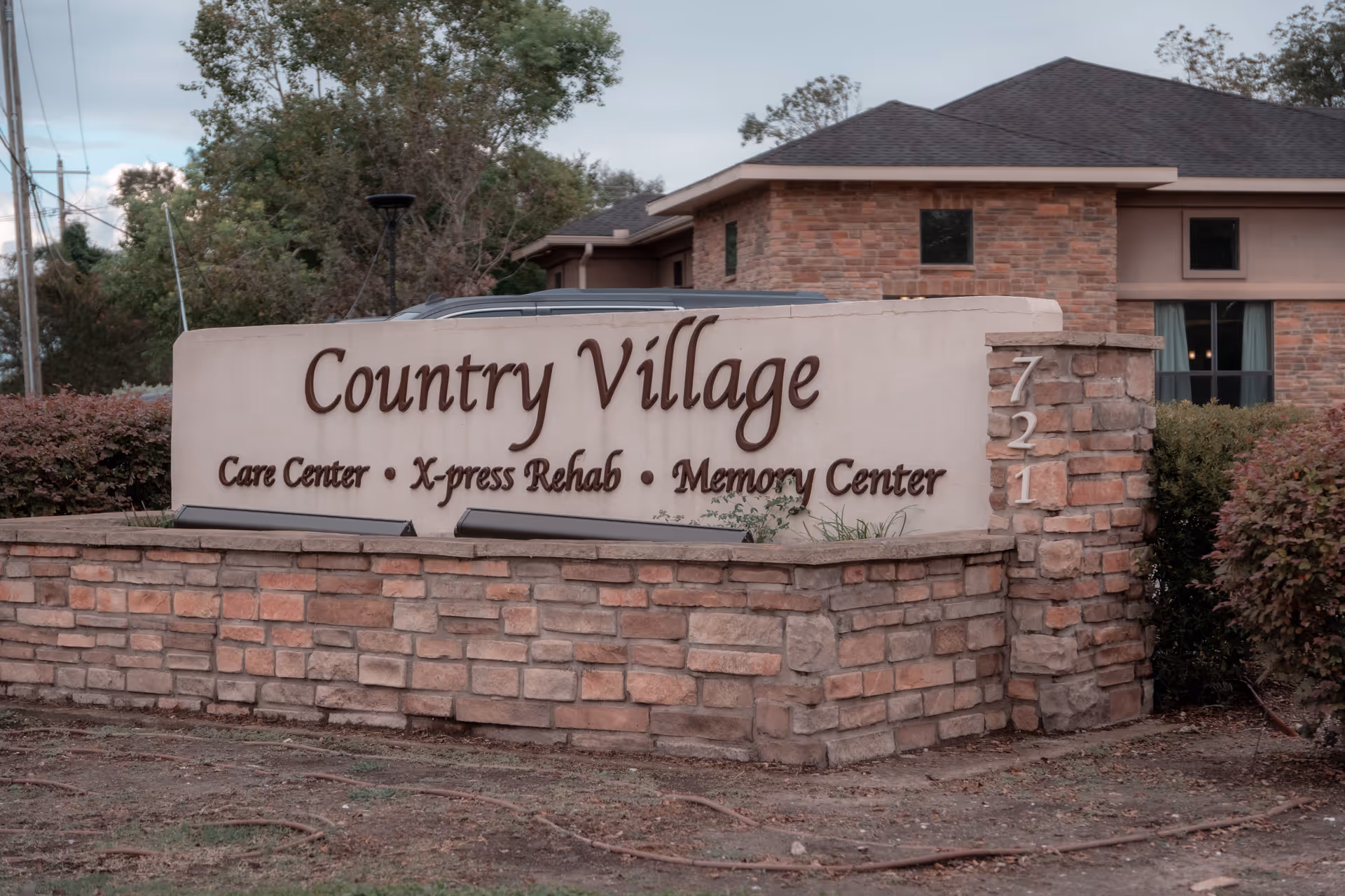 Stone and stucco sign for Country Village Care Center, X-press Rehab, and Memory Center with a brick building and trees in the background.