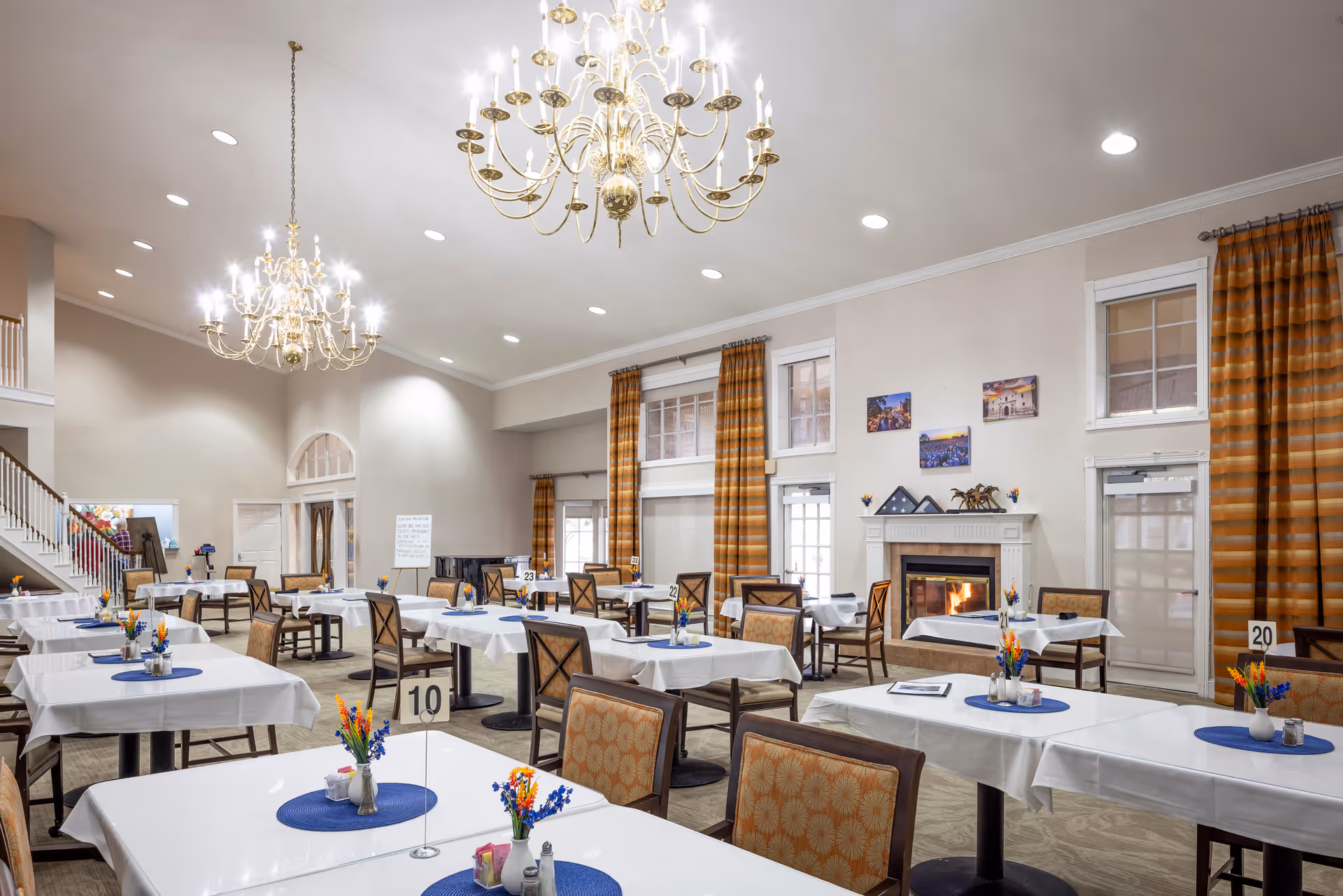 Spacious dining room with white-clothed tables and floral centerpieces, chandeliers overhead and a lit fireplace at the far wall.