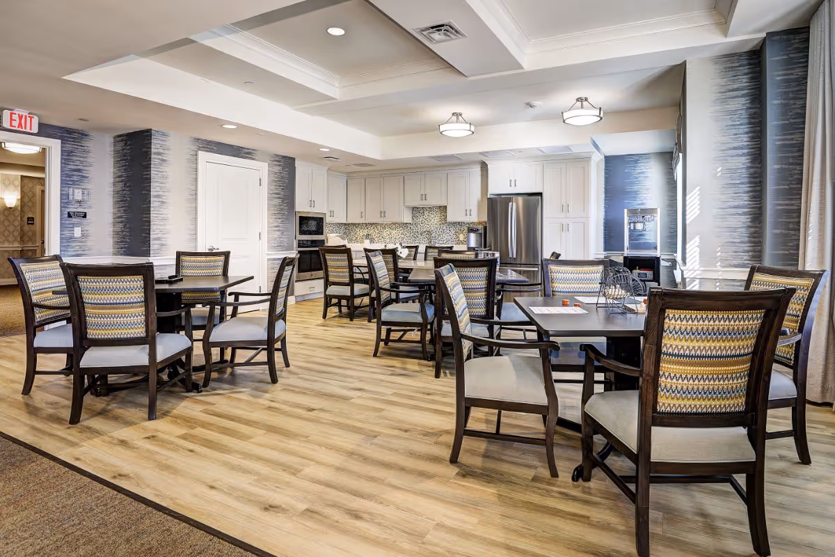 A bright dining area in a senior living facility with several wooden tables and cushioned chairs arranged on a light wood floor. The room features a modern kitchen in the background with white cabinets, a stainless steel refrigerator, and a mosaic tile backsplash. The walls have a blue and white textured pattern, and there is an exit sign above a door on the left side.