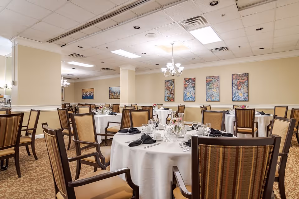 A well-lit dining room with round tables covered in white tablecloths, set with glassware, plates, and black napkins. The room has beige walls adorned with colorful abstract paintings and features wooden chairs with cushioned seats and backs. Ceiling lights and chandeliers provide illumination.