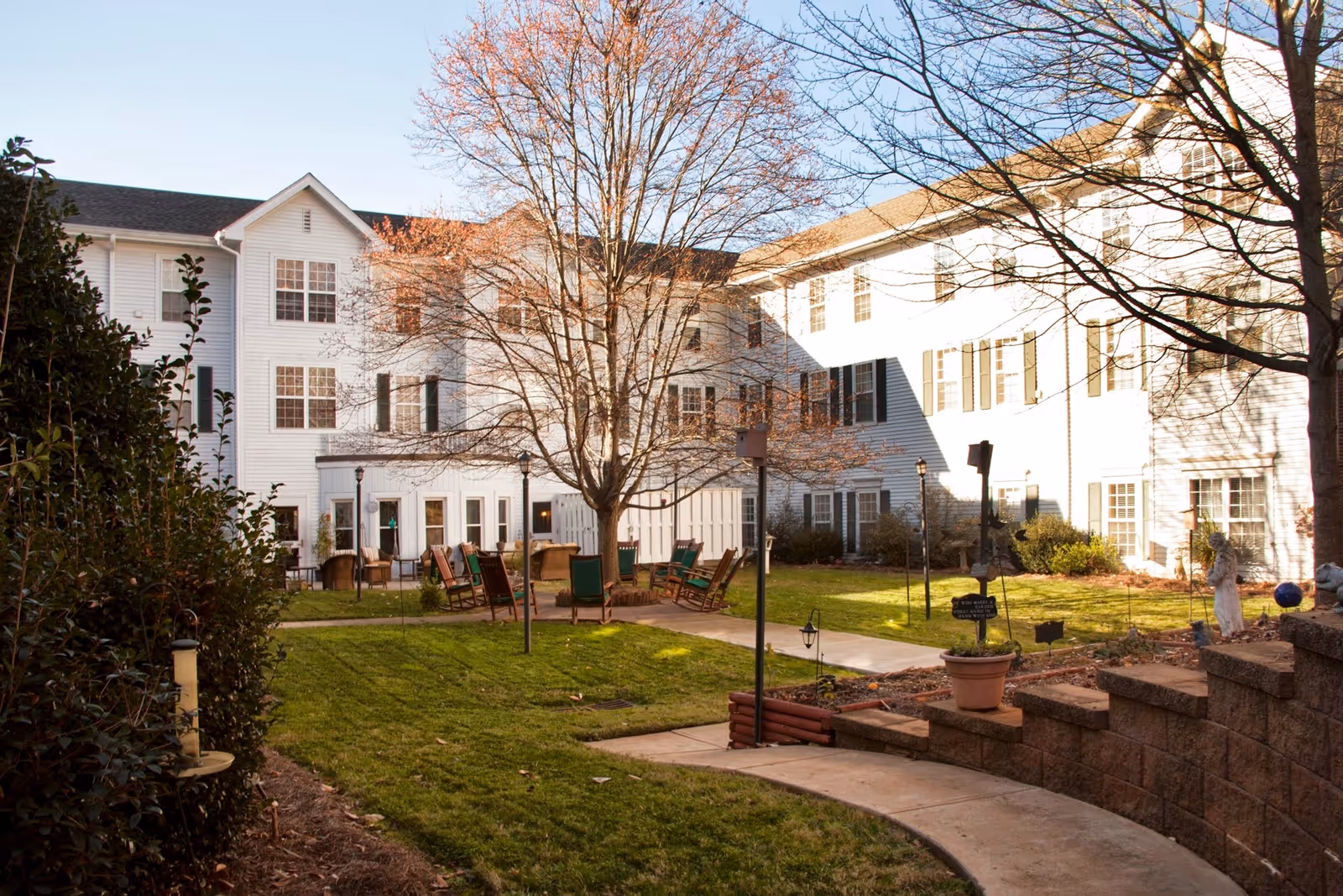 Outdoor courtyard area of a senior living facility with a large tree in the center, surrounded by chairs and tables on a paved patio. The building is white with multiple windows and black shutters. There is a curved walkway leading into the courtyard, green grass, and some landscaping with bushes and potted plants.