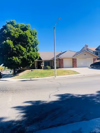 Single-story residential building with a brick and beige exterior, a two-car garage, and a large green tree on the left side. The building is situated on a corner lot with a street and sidewalk in the foreground under a clear blue sky.