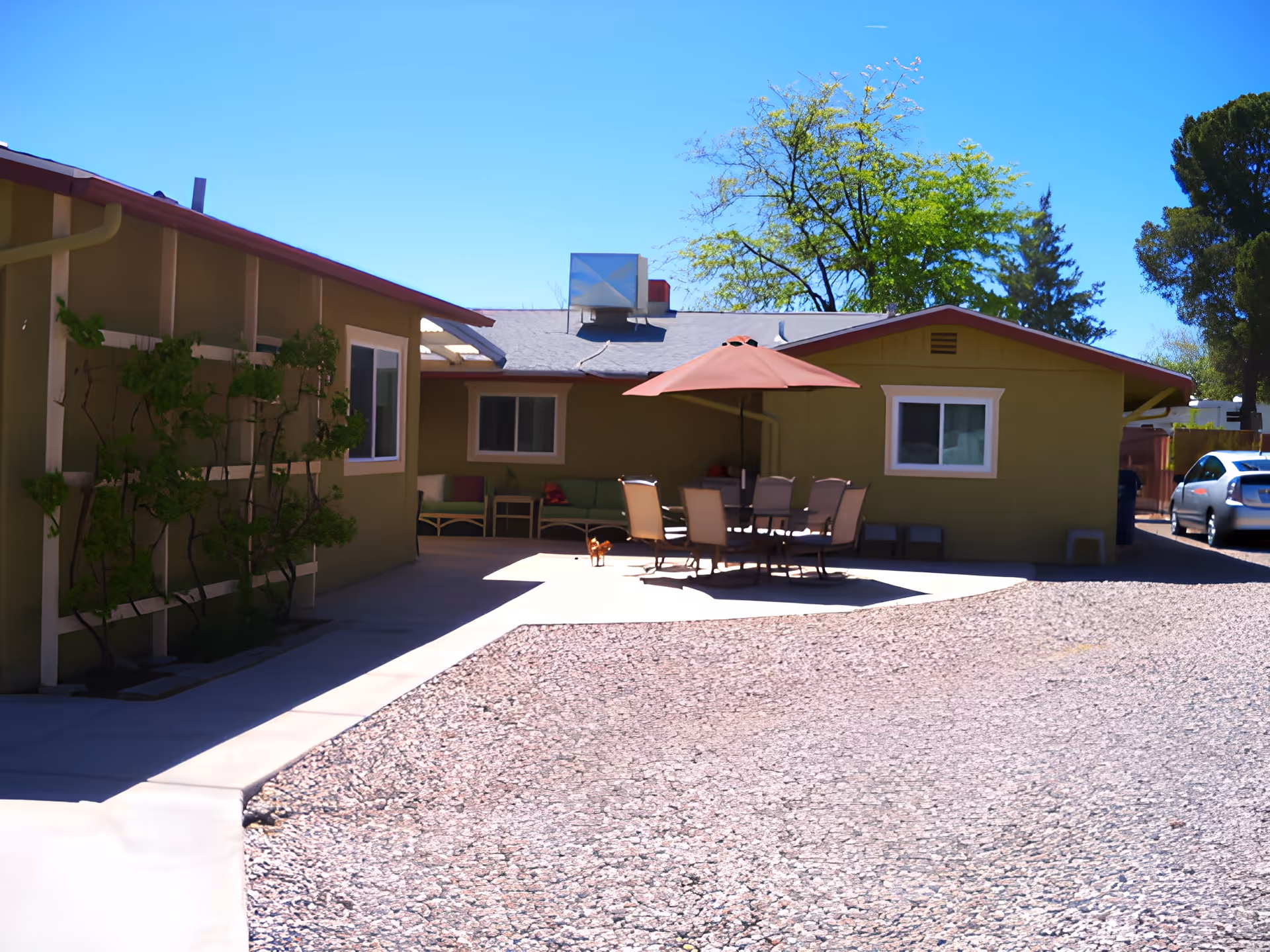 Outdoor courtyard of a single-story building with a patio table, umbrella and chairs in front of a green exterior.