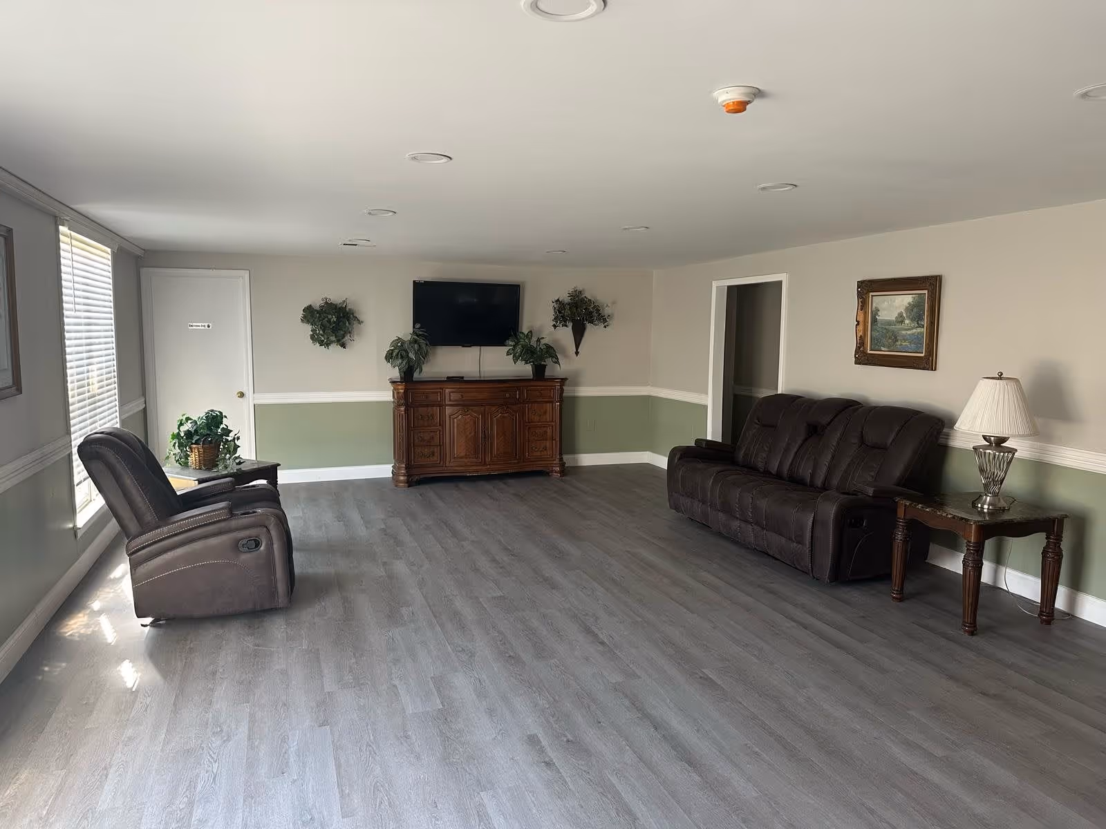 A spacious sitting room with two recliners, a sofa, a TV on a wooden console, and decorative plants and lamps.