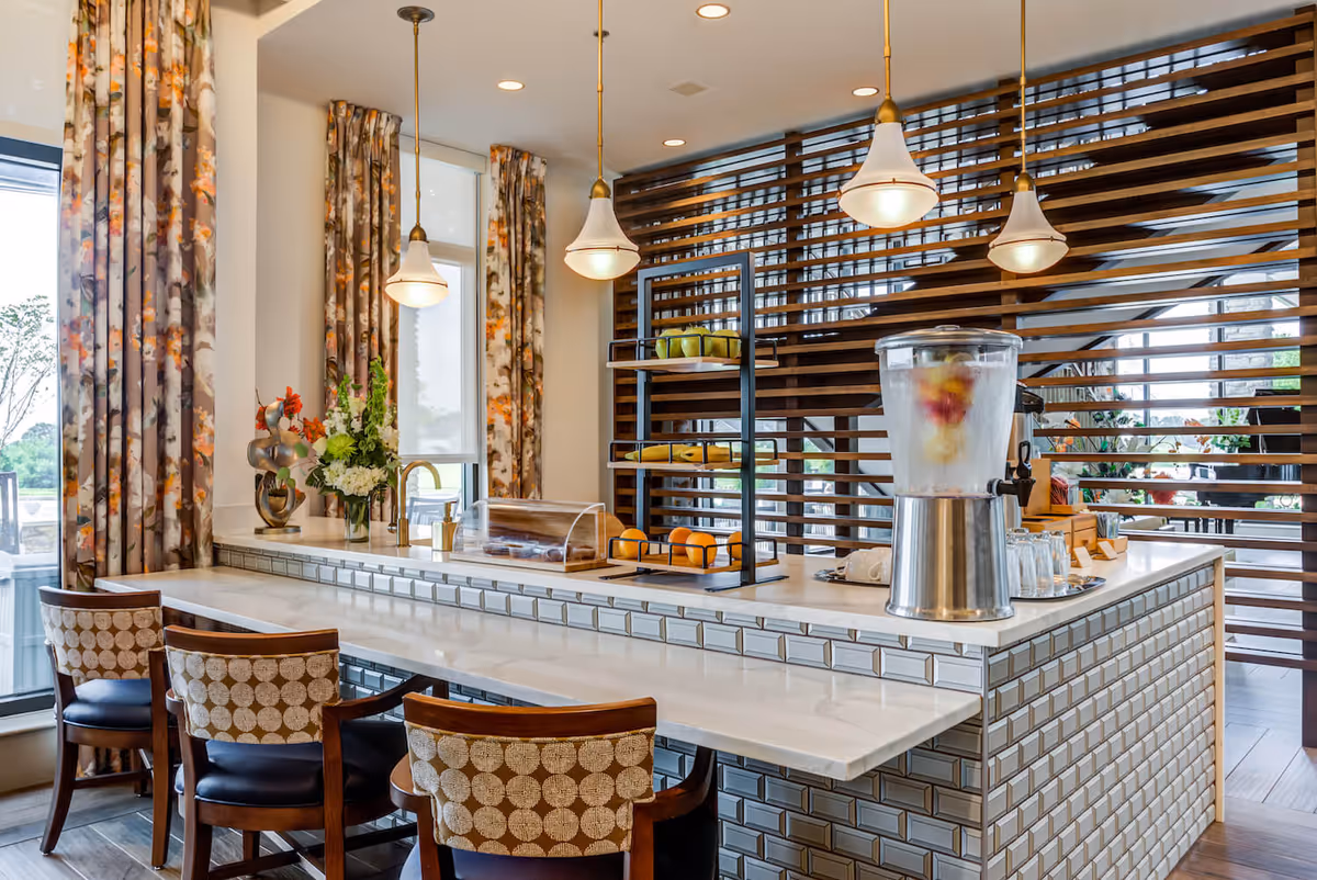 A modern kitchen or dining area with a long white countertop bar featuring three patterned chairs. The counter has a fruit-infused water dispenser, a tray with glasses, a tiered fruit stand with bananas and apples, and a covered pastry tray. The background includes large windows with floral curtains and a decorative wooden slat partition. Three pendant lights hang above the counter.