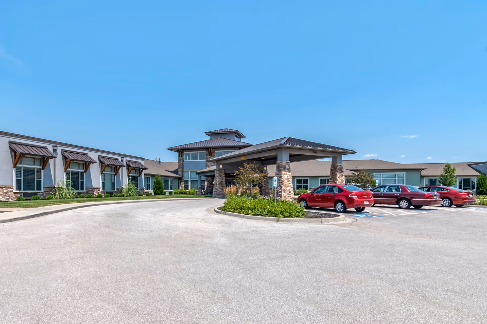 Exterior view of Tiffany Springs Senior Living facility showing a single-story building with a covered entrance supported by stone pillars, several windows with awnings, a parking lot with red cars, and a clear blue sky.