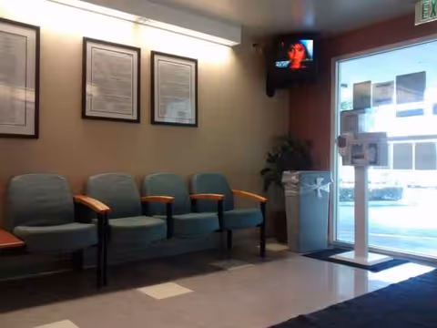 Waiting area with teal chairs, framed documents, plant, and TV near entrance.