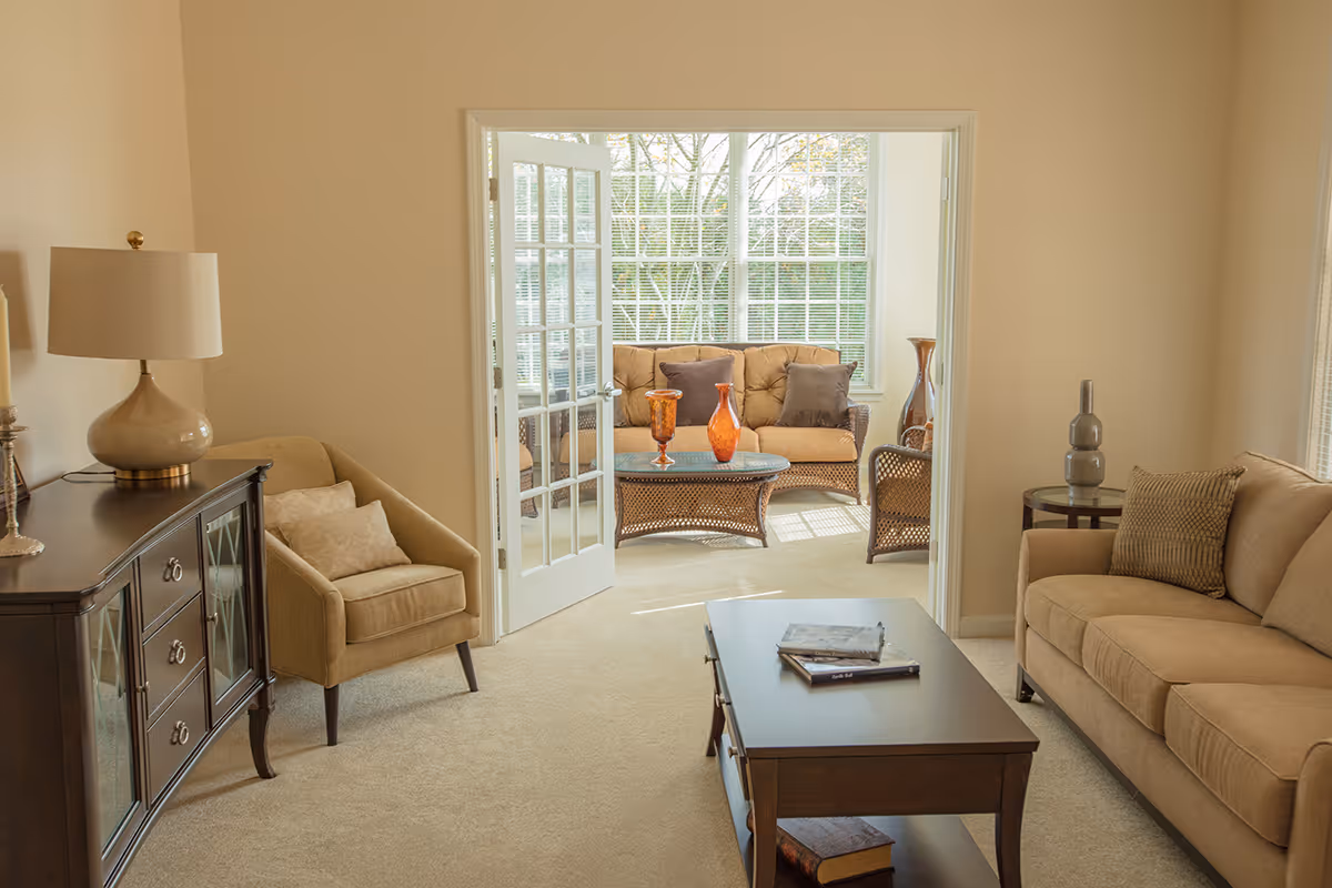 A cozy living room with beige walls and carpet, featuring a beige sofa with a textured pillow, a matching armchair, a dark wooden coffee table with books on it, and a sideboard with a lamp. French doors open to a sunlit sunroom with wicker furniture, including a sofa and chairs, and a glass-top coffee table with decorative orange vases. Large windows in the sunroom reveal green trees outside.