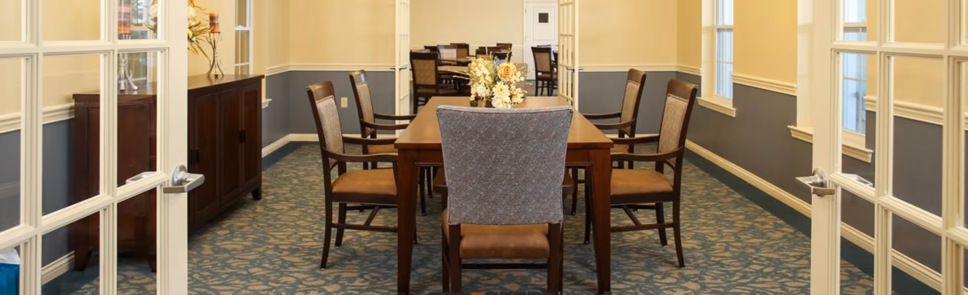 View through double glass doors into a dining room with a long wooden table surrounded by eight chairs. The room has blue and beige walls with white trim, carpeted floor with a floral pattern, and windows on the right side letting in natural light. A sideboard with a flower arrangement is visible on the left wall.