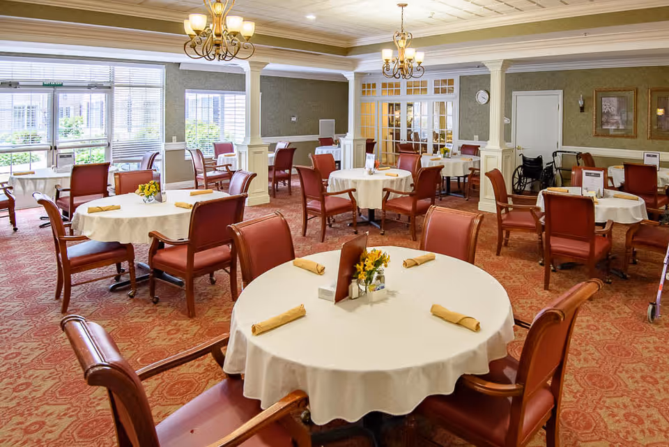 Bright dining room with round tables set with white tablecloths and red chairs in a senior living facility.