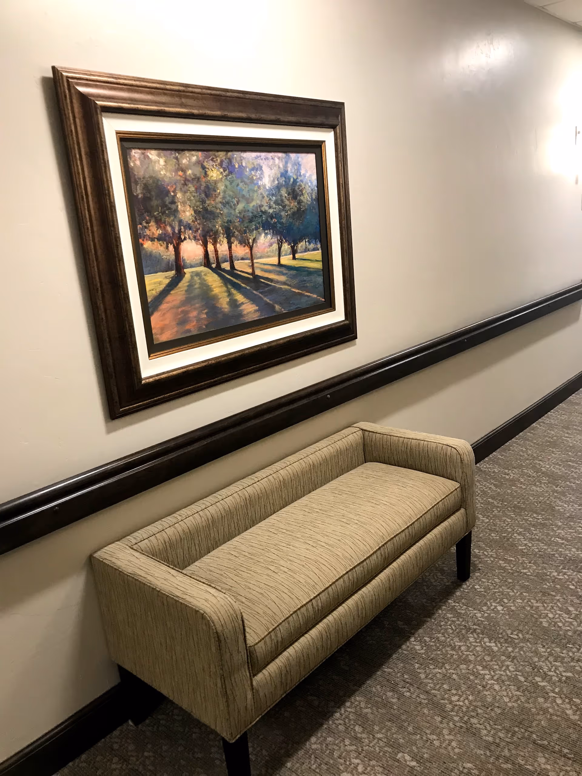 A hallway with a beige upholstered bench against a cream-colored wall. Above the bench hangs a framed painting depicting a row of trees casting long shadows on a grassy area during sunset or sunrise. The hallway has a carpeted floor and a dark wooden chair rail along the wall.