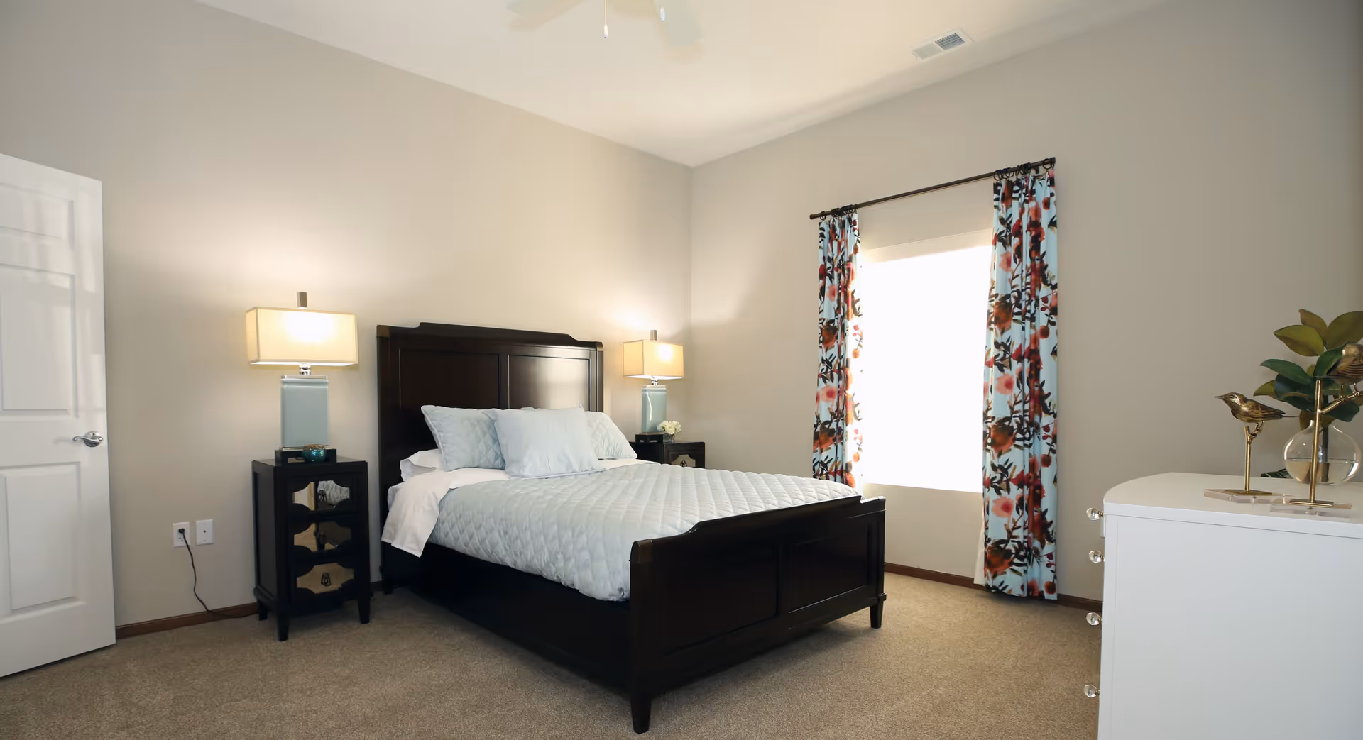 A neatly arranged bedroom with a dark wooden bed frame, light blue bedding, and two matching bedside tables with lamps. There is a window with floral curtains letting in bright natural light, a white dresser with decorative items, and beige carpeted flooring.