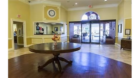 Spacious lobby area with a round wooden table in the center holding a flower arrangement. The room has light yellow walls with white trim, a clock above a reception window, and glass double doors leading outside. There is a hallway to the left and a small table with items on the right side.