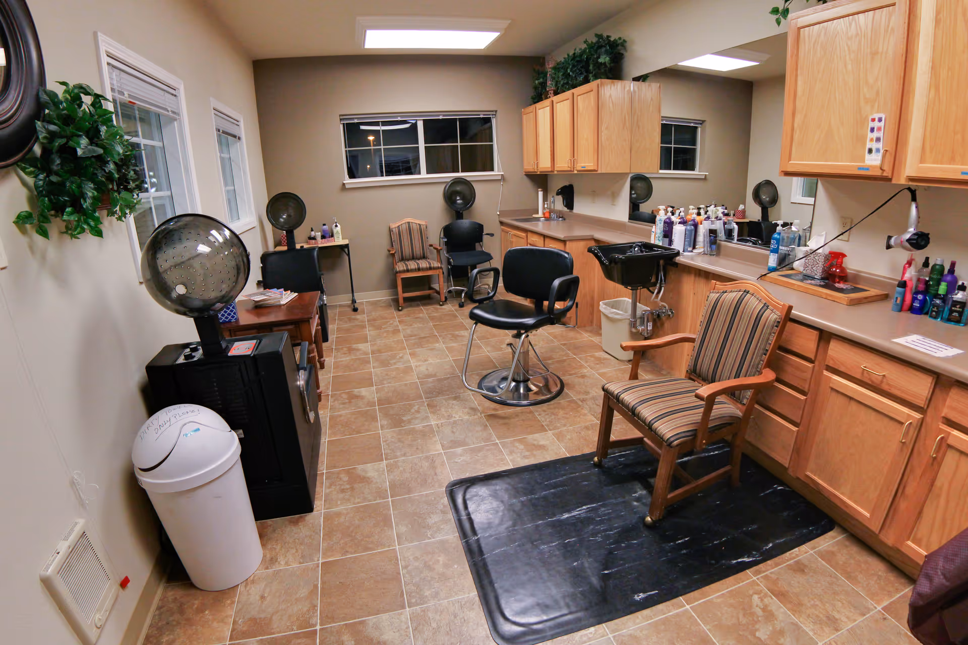 Interior salon room with styling chairs, a shampoo sink, a hooded hair dryer, and countertops lined with hair products and cabinets.