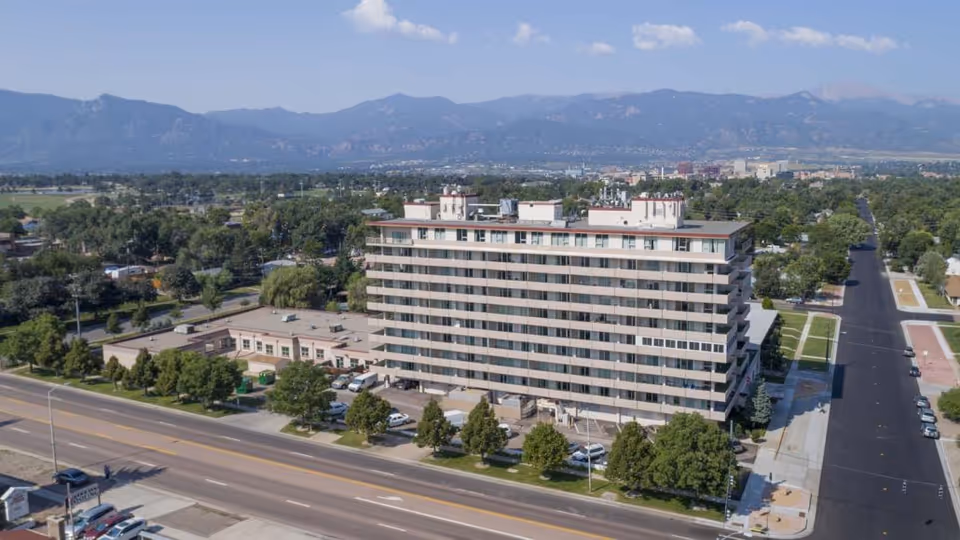 Aerial view of a multi-story building with balconies, surrounded by trees and streets, with mountains visible in the background under a partly cloudy sky.