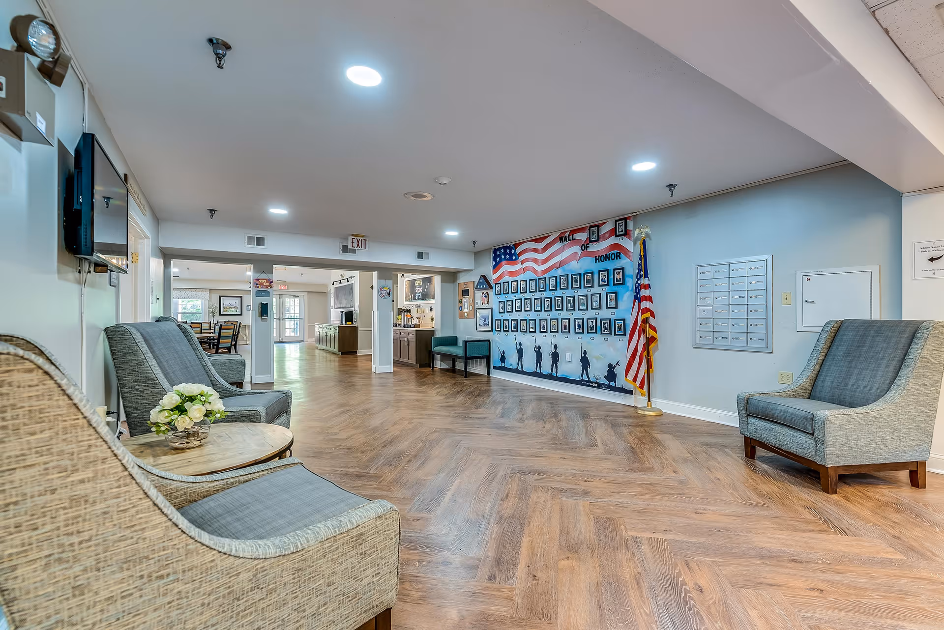 A spacious and well-lit common area in a senior living facility with wooden flooring and several upholstered chairs arranged along the walls. On the right wall, there is a large mural titled 'Hall of Honor' featuring photos and silhouettes of soldiers beneath an American flag, accompanied by a standing American flag. In the background, there is a dining area with tables and chairs and a kitchen or service counter area.
