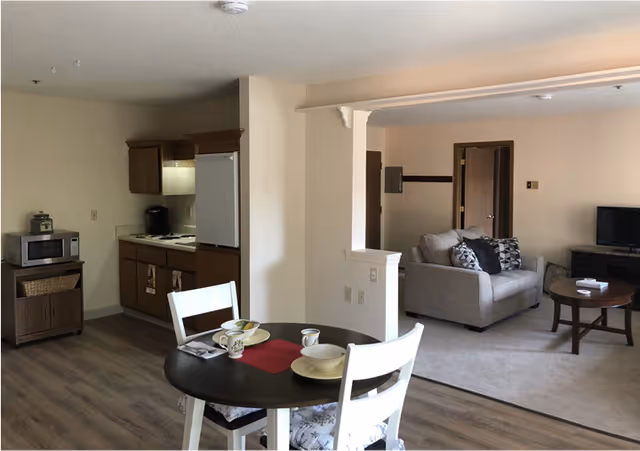 Interior view of a senior living apartment showing a small kitchen area with wooden cabinets, a microwave on a stand, and a refrigerator. In the foreground, there is a round dining table set with plates, cups, and utensils, surrounded by two white chairs. Beyond the dining area, there is a living room with a beige sofa adorned with patterned cushions, a wooden coffee table, and a TV on a stand. The flooring transitions from wood in the kitchen and dining area to carpet in the living room.