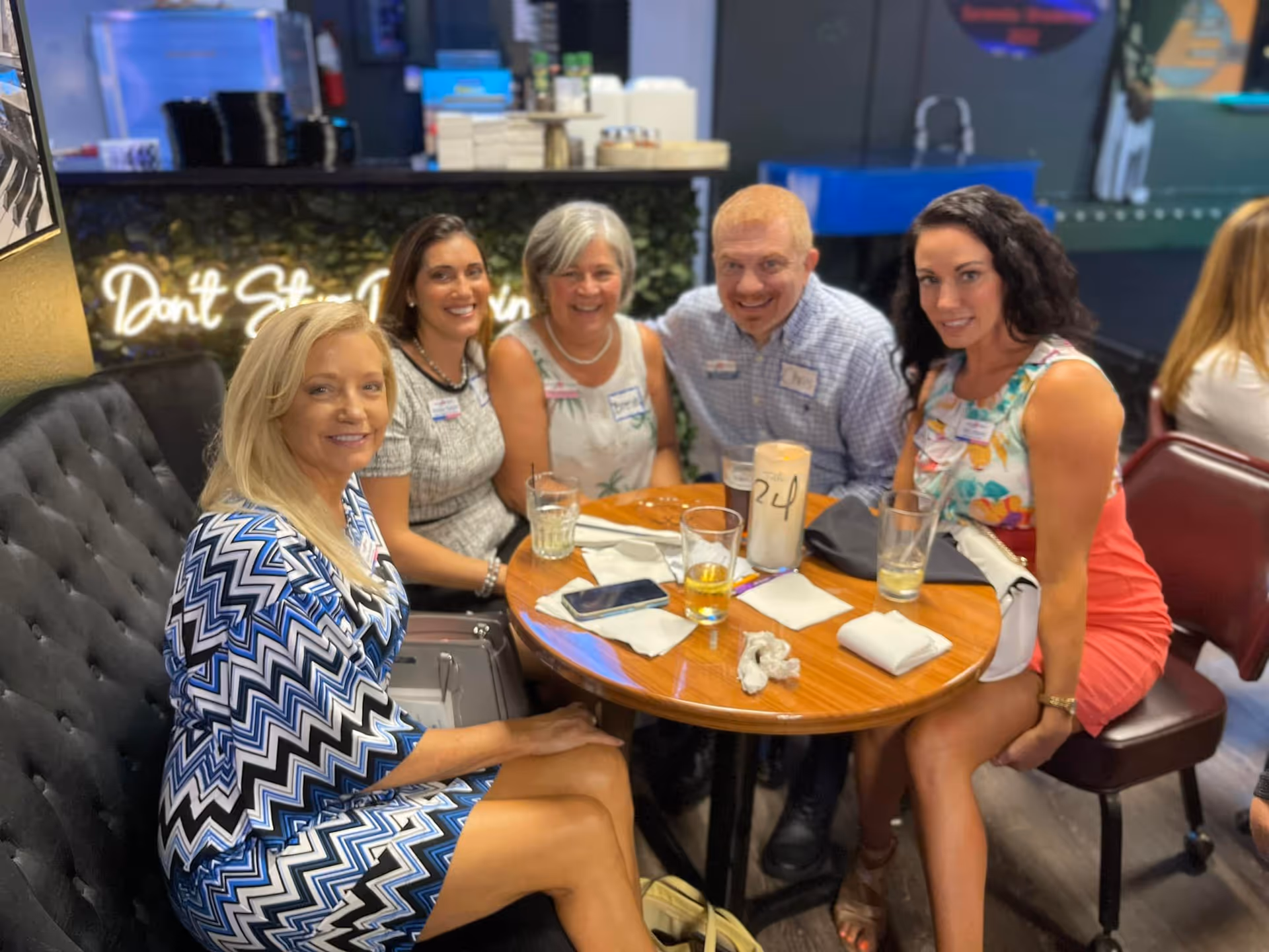 Five adults sitting around a round wooden table in a casual dining or lounge area, smiling at the camera. The table has drinks, napkins, and a phone on it. The background includes a neon sign that says 'Don't Stop' and some shelves with items on them.