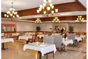 A spacious dining room with multiple tables covered in white tablecloths, surrounded by chairs. The room features chandeliers hanging from a wooden ceiling panel, large windows with curtains, and some potted plants along the walls. A few people are seated at the tables.