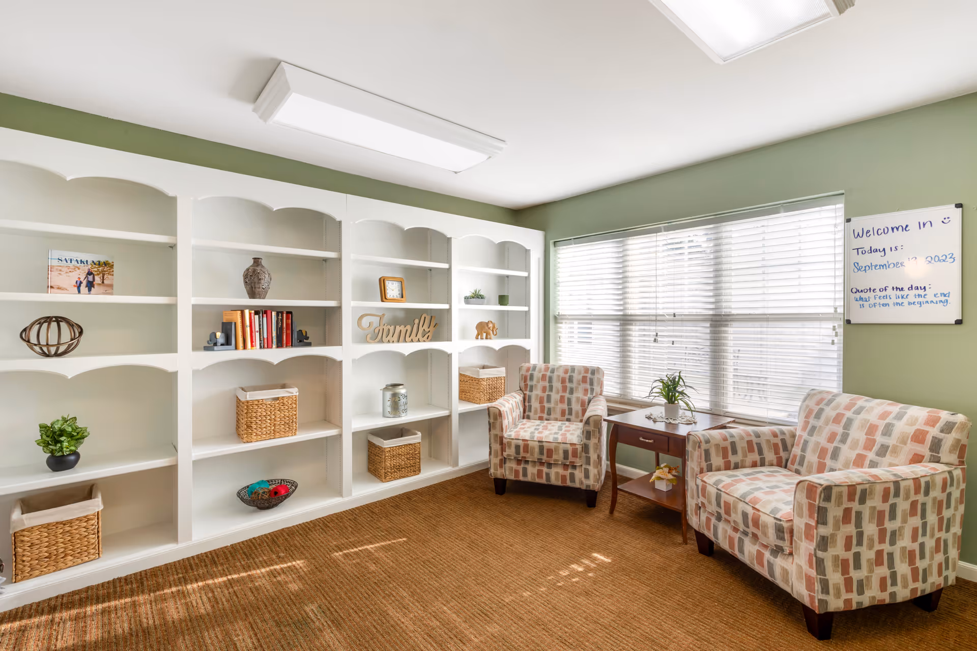 A cozy sitting area with two patterned armchairs and a small wooden side table with a plant on it, positioned in front of a large window with white blinds. To the left, there is a white built-in bookshelf with various decorative items including books, baskets, a clock, and a wooden 'Family' sign. The walls are painted green and there is a whiteboard on the wall with a welcome message and the date September 13, 2023.