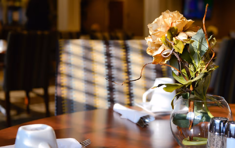 Close-up of a dining table set with white napkins, silverware, and a glass vase holding a bouquet of light-colored flowers. In the background, there are patterned upholstered chairs and a softly lit dining area.